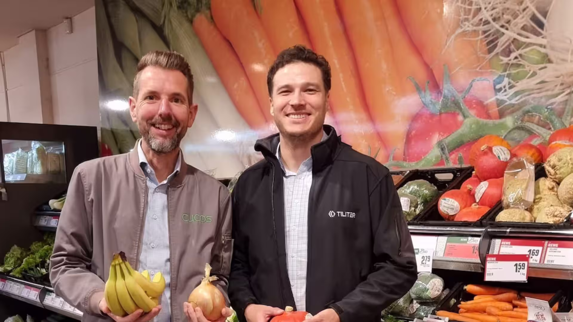 Two colleagues standing together in a supermarket produce aisle, one holding a bunch of bananas
