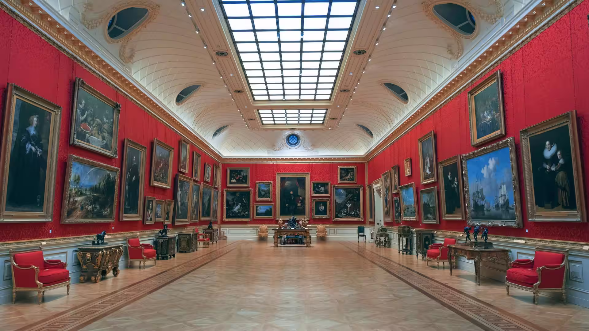 A museum gallery hung with framed paintings on deep red walls