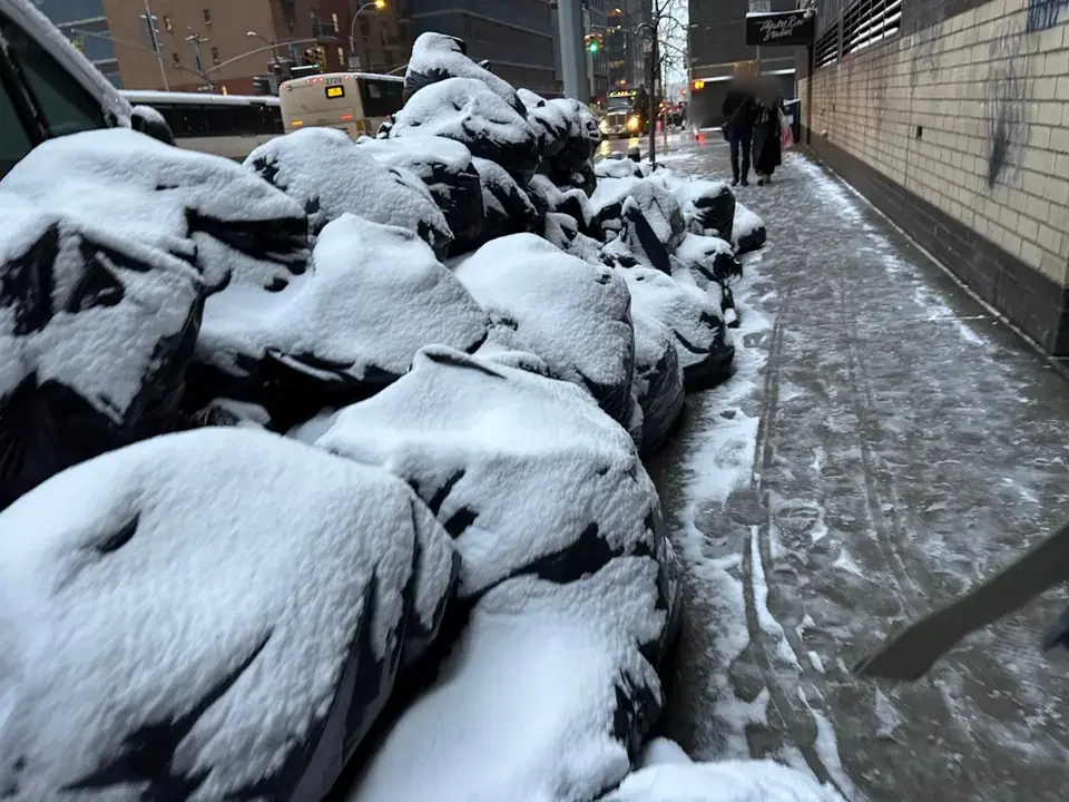 Snow-covered rubbish bags piled beside an icy footpath